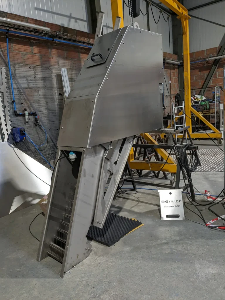 Vue trois-quarts du dégrilleur escalier BioScreen DGE Biotrade suspendu au pont roulant en atelier, avec panneau Biotrade au sol